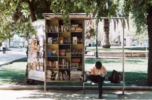 Man on bench reading book next to public mini-library outside
