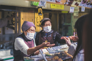 Two Asian women exchanging food and money at an outdoor street market while wearing masks